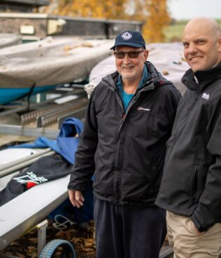 An image of commadore John from ulley Sailing club and Tom chaplin from onpath energy, standing next to some of the new boats Ulley sailing club has purchased with community fund grants